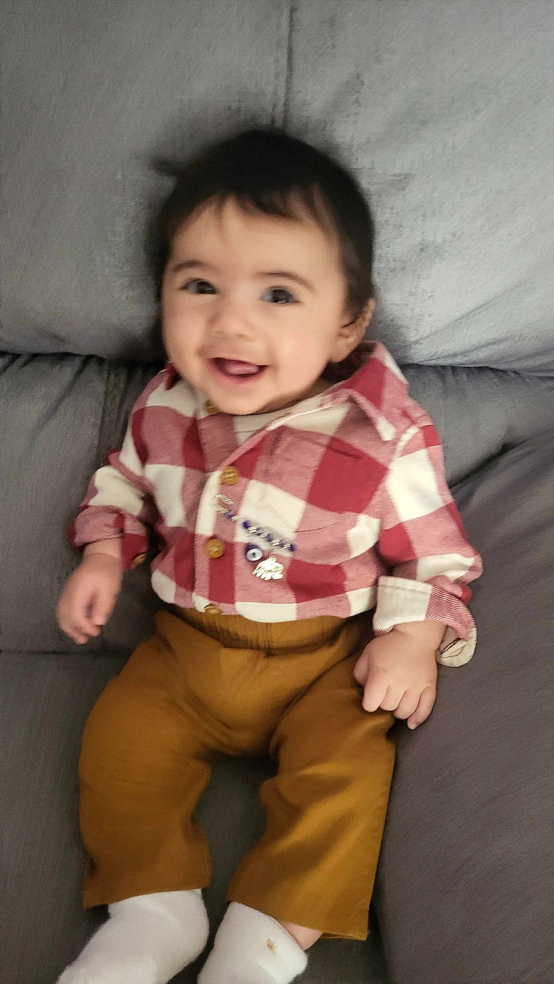 Sara is registered to the contest to win money with this photo: baby, child, smiling, red_shirt, checkered_shirt, mustard_pants, white_socks, couch, gray_couch, happy, indoor, portrait, infant, clothing, sitting, cute, young_child, face, person, casual