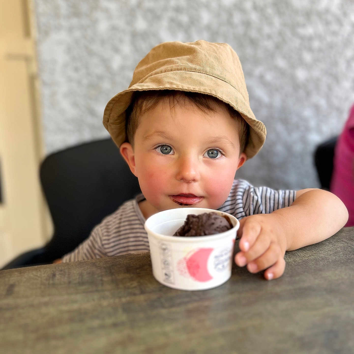 Xuann a rejoint le concours — aidez-le/la à gagner de superbes lots ! bucket_hat, child, chocolate_ice_cream, cup, cute, dessert, eyes, face, food, hand, hat, indoor, messy, person, portrait, smile, striped_shirt, sweet, table, toddler
