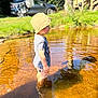 child, water, pond, hat, striped_shirt, shorts, sandals, reflection, ripples, sunlight, grass, car, clothes_drying_rack, tree, outdoor, summer, nature, daylight, backyard, person