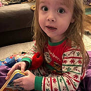 Delilah is registered to the contest to win money with this photo: child, pajamas, toy, book, indoor, living_room, blanket, curly_hair, sitting, colorful, cute, holiday_theme, holding, person, young_child, playtime, cozy, smiling, furniture, home