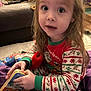 child, pajamas, toy, book, indoor, living_room, blanket, curly_hair, sitting, colorful, cute, holiday_theme, holding, person, young_child, playtime, cozy, smiling, furniture, home