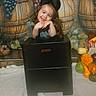 barrels, black_box, blue_dress, bow, child, costume, face, farm, fruit, girl, grapes, hands, harvest, indoor, long_hair, plush_toy, portrait, pumpkins, smiling, vegetables