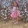 agriculture, child, cotton_bolls, cotton_field, daylight, fashion, girl, hair_bow, long_hair, nature, outdoor, pink_dress, pink_skirt, plants, portrait, shoes, smiling, socks, standing, toddler