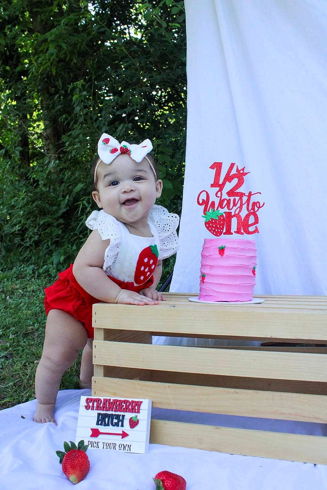Leilani joined the competition — help win amazing prizes! baby, child, cake, pink_cake, strawberry, strawberry_patch, outdoor, greenery, wooden_crate, white_cloth, headband, smiling, celebration, half_birthday, food, sign, person, grass, nature, cute
