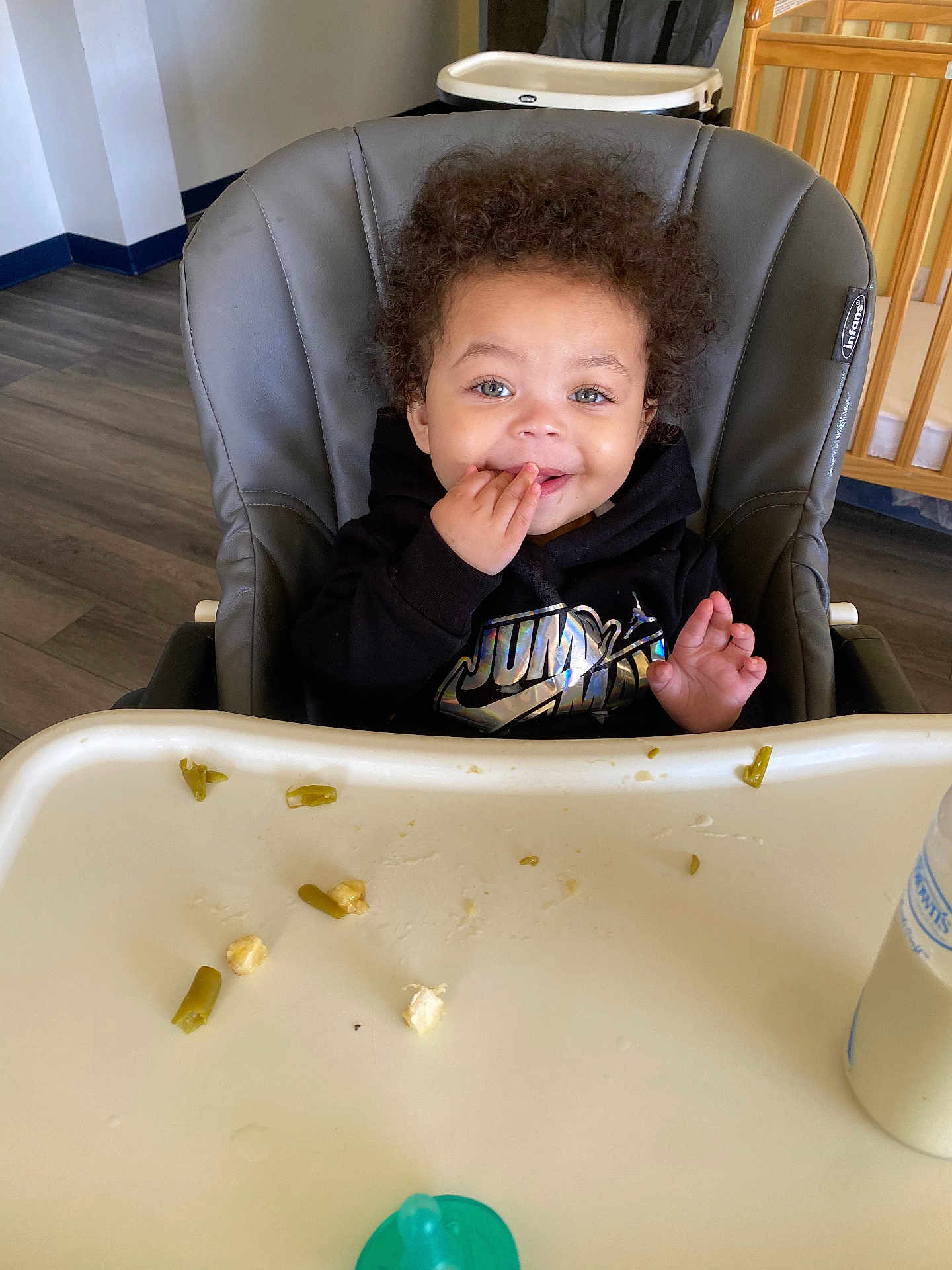 Trenton is registered to the contest to win money with this photo: toddler, child, high_chair, curly_hair, black_hoodie, food, tray, baby_bottle, indoor, flooring, crib, wooden_furniture, happy, eating, smiling, person, face, hand, home, casual