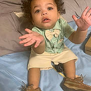 Trenton is registered to the contest to win money with this photo: toddler, child, boy, curly_hair, shirt, bow_tie, shorts, brown_shoes, bed, pillow, blanket, indoor, person, expression, hands, open_mouth, sitting, looking_up, cute, clothing