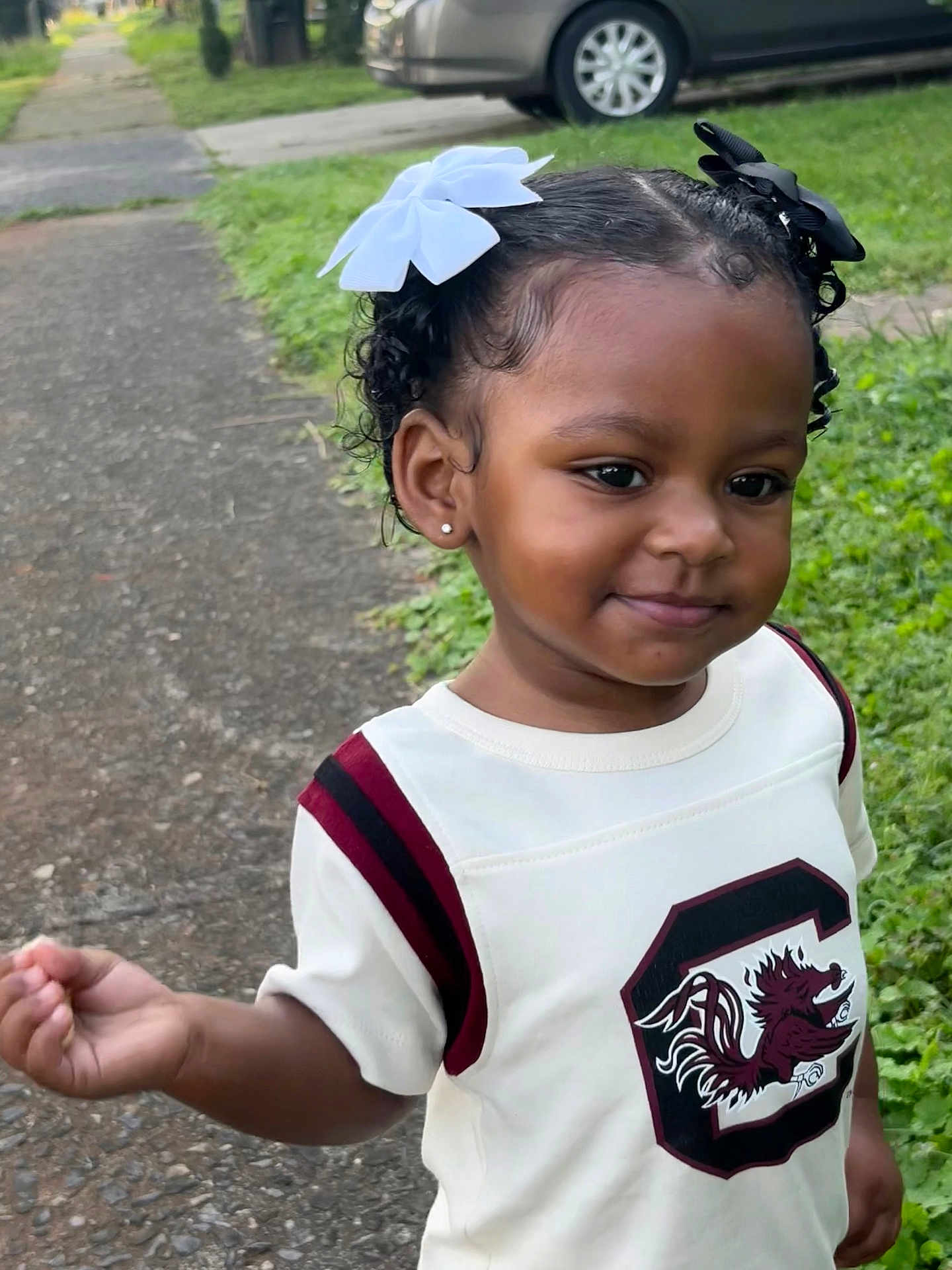 Khiari Easley is registered to the contest to win money with this photo: child, toddler, outdoor, sidewalk, grass, car, white_shirt, hair_bows, curly_hair, smiling, earrings, walking, young_child, greenery, casual_clothing, portrait, daylight, happy, person, cute