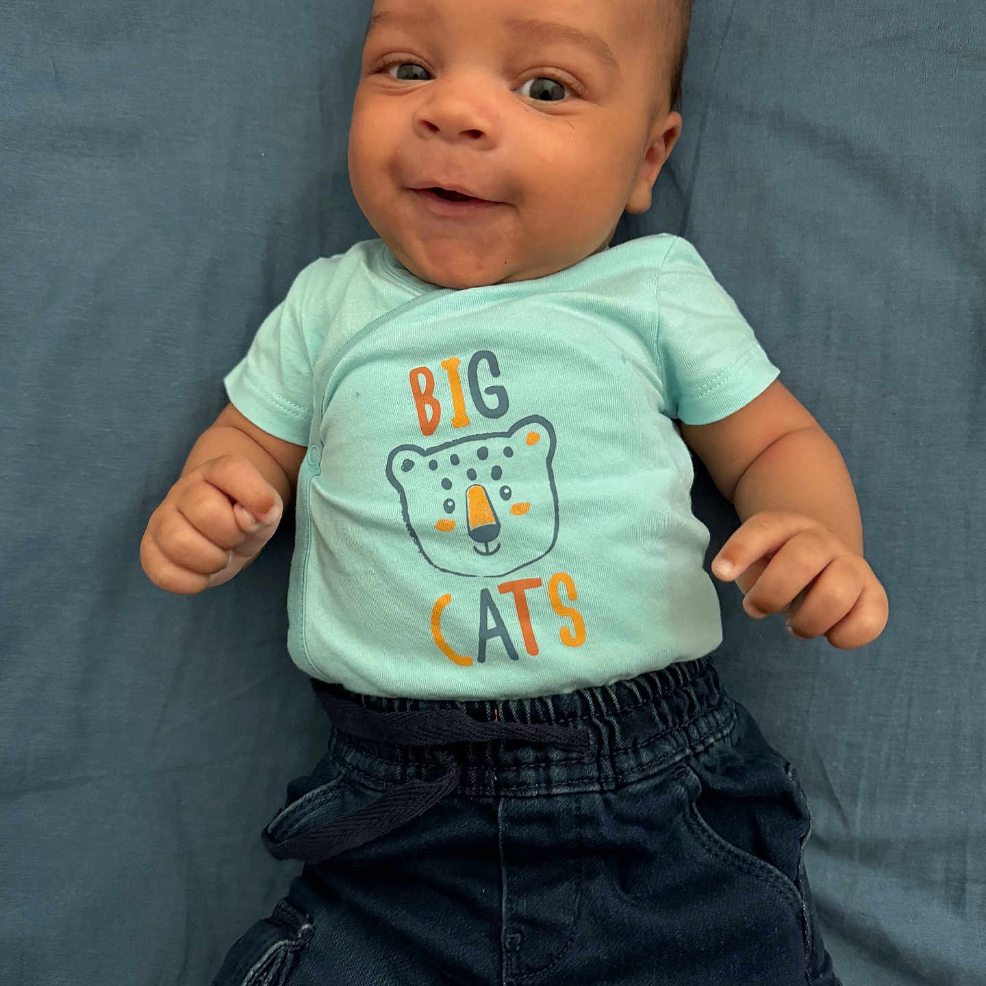 Saël a rejoint le concours — aidez-le/la à gagner de superbes lots ! baby, big_cats, blanket, blue_shirt, cargo_pants, casual_clothing, child, closeup, cute, dark_pants, fists, graphic_tee, happy, indoors, infant, lying_down, person, portrait, smiling, young_child