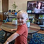 Hunter is registered to the contest to win money with this photo: toddler, child, indoor, living_room, plant, table, television, carpet, furniture, person, smiling, blond_hair, onesie, wood_floor, decor, home, couch, pot, screen, playful