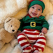 Fabio a rejoint le concours — aidez-le/la à gagner de superbes lots ! baby, infant, christmas_elf_costume, striped_clothing, teddy_bear, stuffed_animal, smile, happy, cute, holiday, festive, plush, soft_texture, blanket, cozy, child, portrait, indoors, cute_outfit, hat