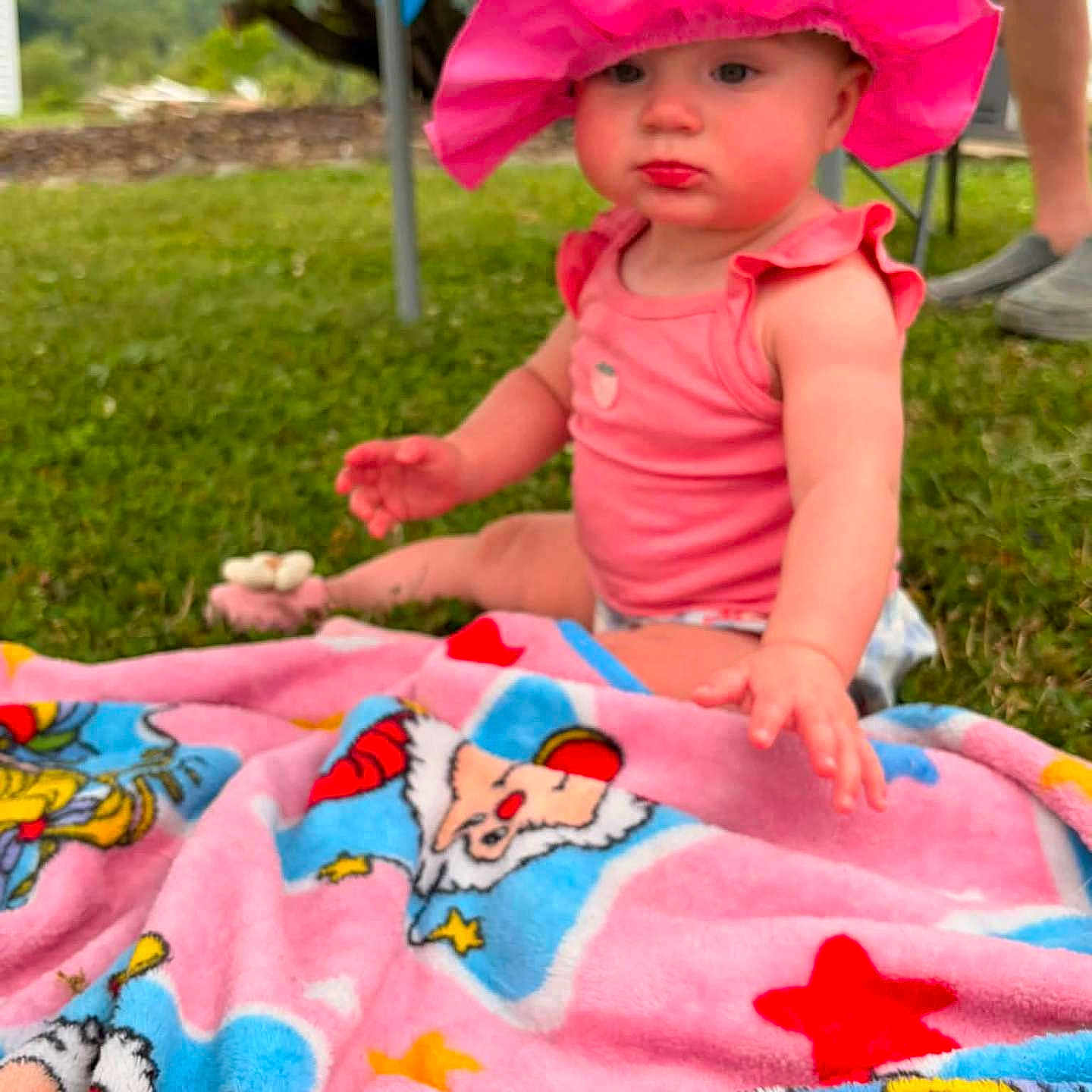 Judith joined the competition — help win amazing prizes! baby, child, pink_hat, cow_print, blanket, outdoor, grass, colorful, cartoon, clothing, sitting, person, summer, sunhat, cute, pout, greenery, legs, footwear, nature