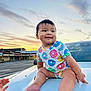 baby, child, infant, car, hood, sunset, outdoor, clothing, colorful, donut_pattern, smile, happy, cute, hands, feet, sky, clouds, road, urban, parking_lot