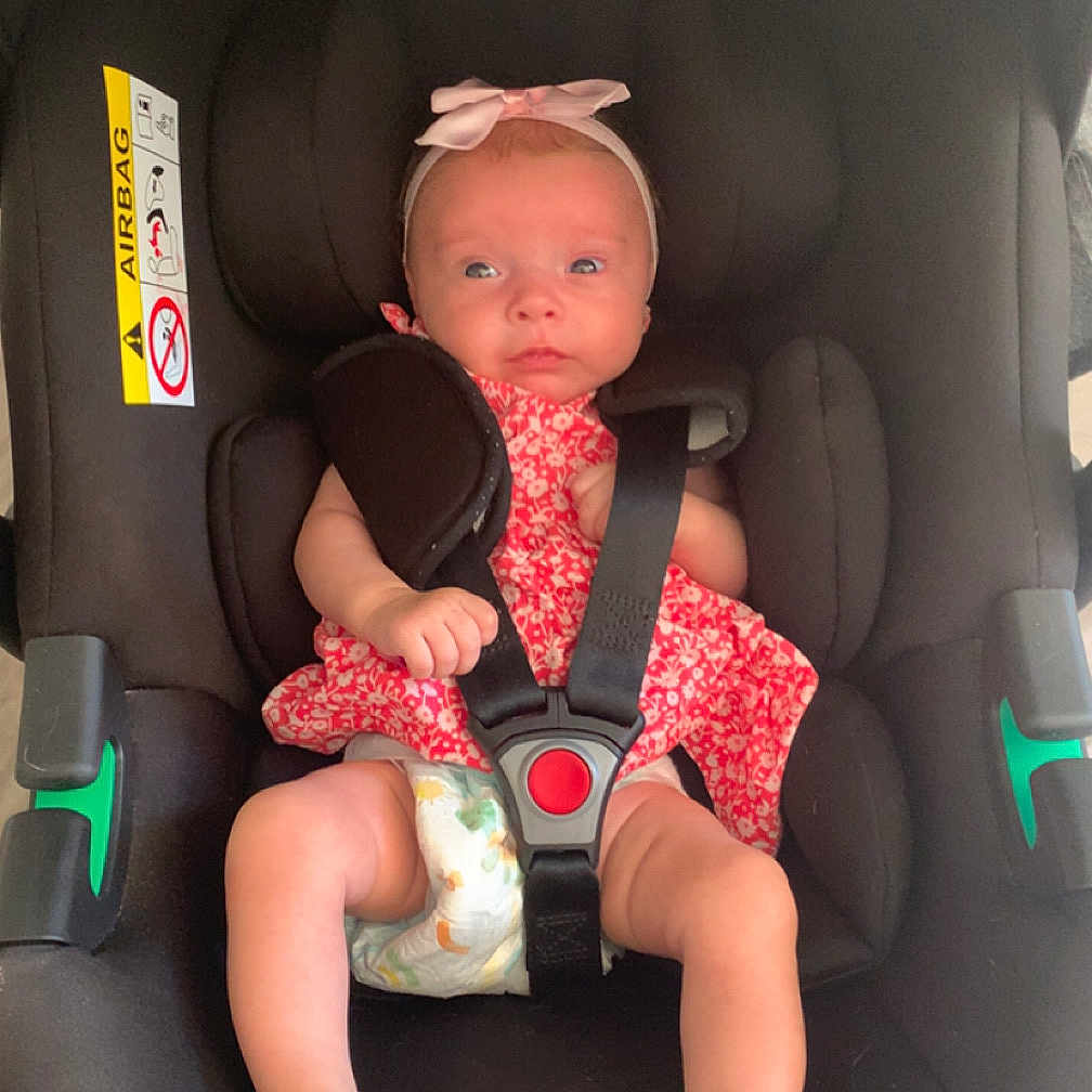 Alessia a rejoint le concours — aidez-le/la à gagner de superbes lots ! baby, baby_girl, black_cushion, car_seat, child, curious, cute, diaper, floral_dress, harness, headband, indoor, infant, newborn, person, pink_bow, red_button, seatbelt, sitting, small_feet