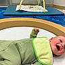 Mateo is registered to the contest to win money with this photo: baby, infant, smiling, green_onesie, bib, play_mat, indoor, child, cute, happy, floor, toy, blue_mat, padded, cheeks, lying_down, room, play_area, young_child, face