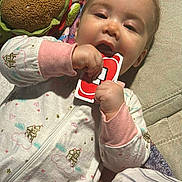 Amelia is registered to the contest to win money with this photo: baby, child, pajamas, plush_toy, toy, uno_card, hand, face, couch, blanket, paper, indoor, clothing, infant, playing, lying_down, cute, chewing, soft_toy, relaxed