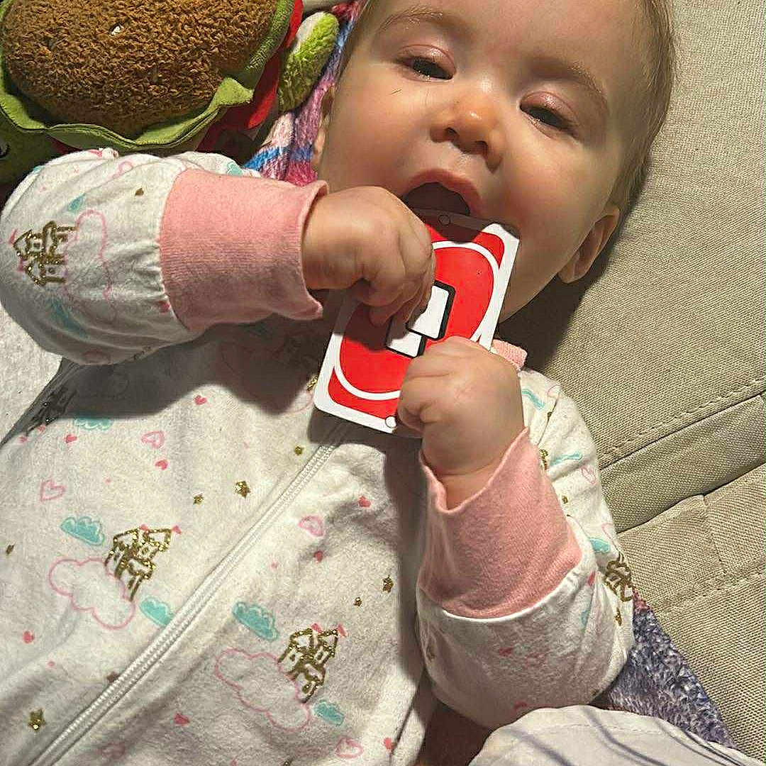 Amelia is registered to the contest to win money with this photo: baby, blanket, chewing, child, clothing, couch, cute, face, hand, indoor, infant, lying_down, pajamas, paper, playing, plush_toy, relaxed, soft_toy, toy, uno_card