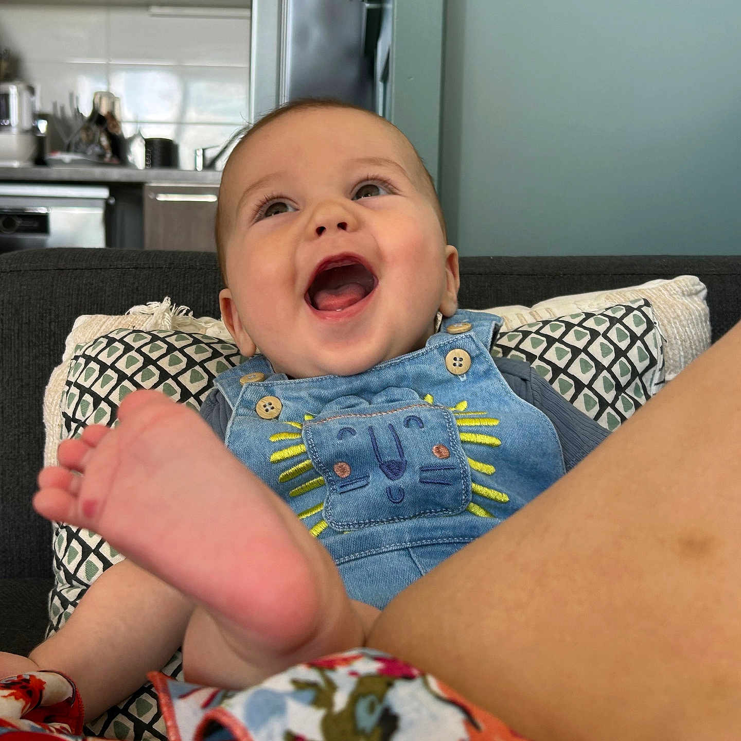 Elio a rejoint le concours — aidez-le/la à gagner de superbes lots ! baby, child, clothing, colorful, couch, cushions, denim_overalls, expression, fabric, face, foot, happy, home_interior, infant, kitchen, lion_patch, person, portrait, sitting, smiling