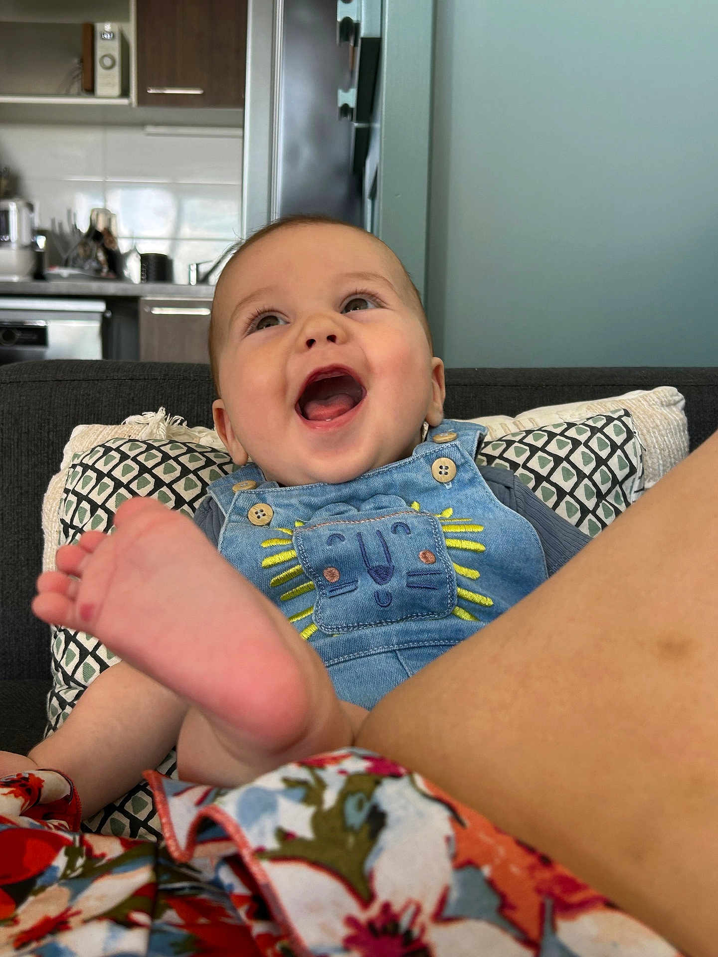Elio a rejoint le concours — aidez-le/la à gagner de superbes lots ! baby, child, smiling, denim_overalls, lion_patch, foot, couch, cushions, kitchen, home_interior, happy, infant, person, portrait, clothing, fabric, colorful, sitting, face, expression