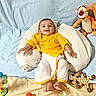 baby, child, yellow_clothing, pillow, blue_backdrop, toys, stuffed_tiger, giraffe_toy, broccoli, food_bowl, smiling, happy, indoor, playful, decorations, heart_sticker, moon_sticker, infant, cute, cozy