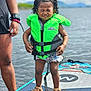 child, life_jacket, water, paddleboard, outdoor, summer, sandals, water_sports, person, adult, lake, nature, recreation, safety, happy, standing, curly_hair, water_sports_equipment, shorts, sunlight