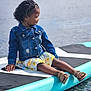 child, denim_jacket, curly_hair, paddleboard, water, sandals, outdoor, smiling, sitting, young_child, nature, lake, summer, relaxing, casual_clothing, floral_skirt, calm, side_view, peaceful, portrait
