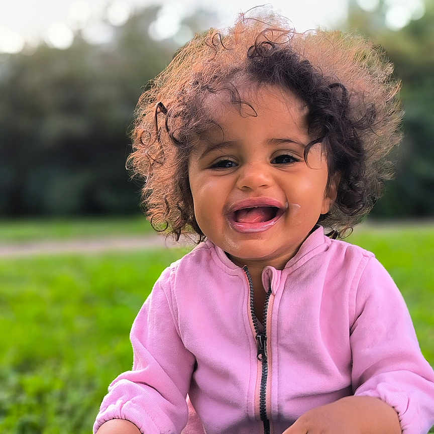 Mona participe au concours pour gagner de l'argent avec cette photo : casual_clothing, child, curly_hair, cute, fun, grass, greenery, happy, jeans, lap, laughing, nature, outdoor, person, pink_jacket, portrait, smiling, sunlight, toddler, young_child