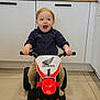 toddler, child, toy, motorbike, indoor, kitchen, floor, happy, excited, vehicle, small, person, playing, clothing, pants, shirt, footwear, window, cabinet, handlebars