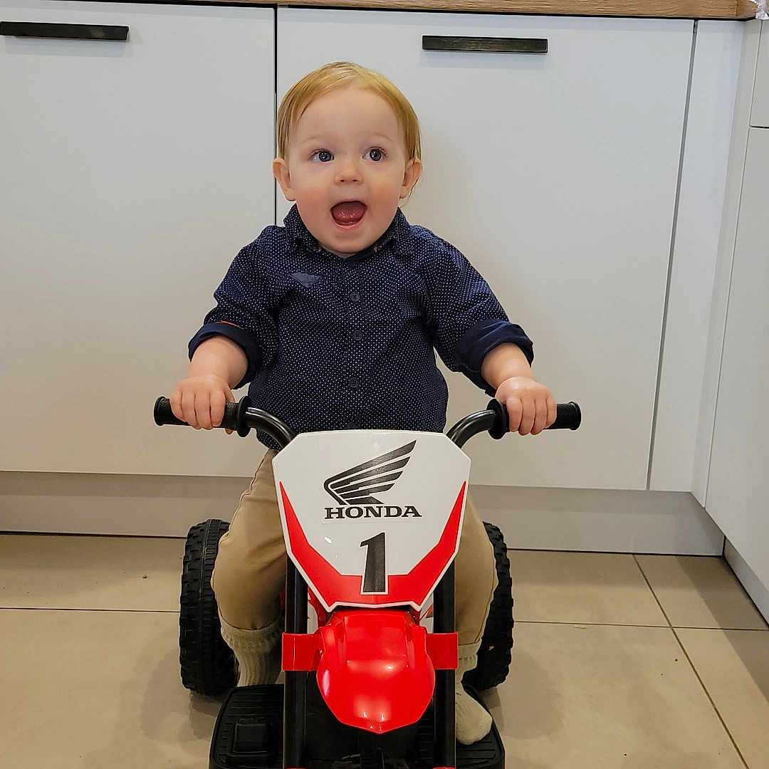 Gabriel a rejoint le concours — aidez-le/la à gagner de superbes lots ! cabinet, child, clothing, excited, floor, footwear, handlebars, happy, indoor, kitchen, motorbike, pants, person, playing, shirt, small, toddler, toy, vehicle, window