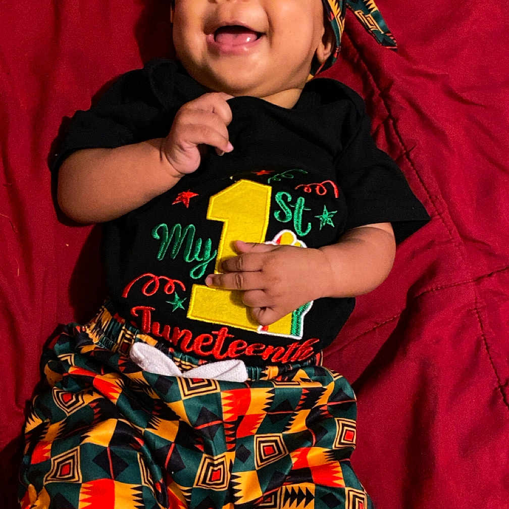Jaiceon joined the competition — help win amazing prizes! baby, child, smiling, happy, portrait, lying_down, red_blanket, headwrap, patterned_clothing, colorful, geometric_pattern, black_shirt, pants, infant, face, hands, indoors, fashion, text_on_clothing, closeup