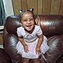 child, toddler, smiling, happy, dress, unicorn, pink, hair_buns, leather_chair, armchair, wood_paneling, indoor, cute, person, fun, joy, small_child, portrait, clothing, furniture