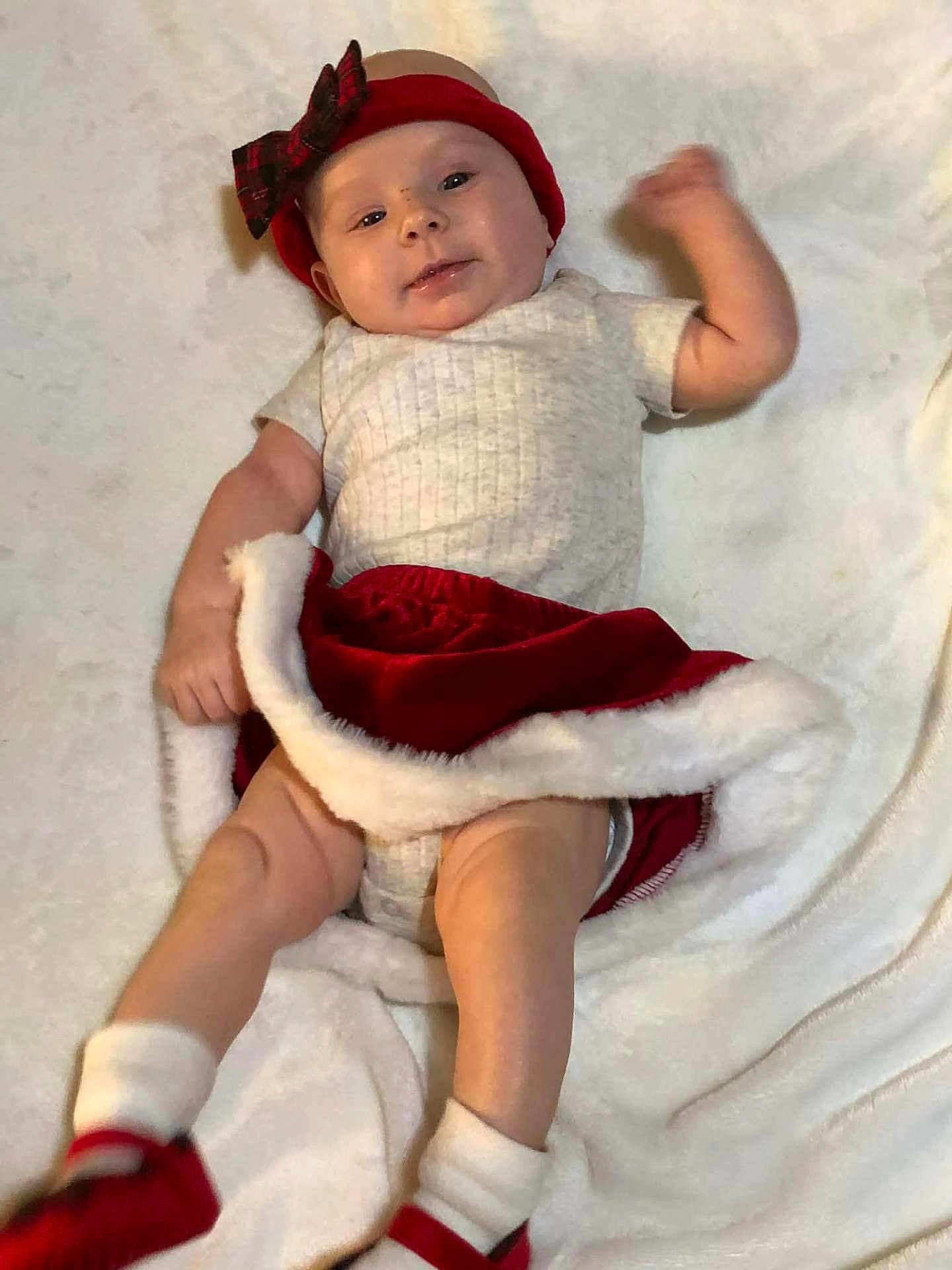 Emberly is registered to the contest to win money with this photo: baby, infant, smile, headband, bow, red_skirt, white_onesie, socks, red_shoes, blanket, lying_down, cute, happy, holiday, festive, child, young, soft_texture, indoors, portrait