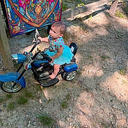 Eliana joined the competition — help win amazing prizes! toddler, child, toy_motorcycle, outdoor, dirt_path, sunlight, shadow, blue_tutu, colorful_tapestry, grass, summer, playtime, cute, young_child, small_vehicle, nature, sunny_day, barefoot, curious, portrait