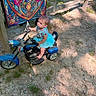 toddler, child, toy_motorcycle, outdoor, dirt_path, sunlight, shadow, blue_tutu, colorful_tapestry, grass, summer, playtime, cute, young_child, small_vehicle, nature, sunny_day, barefoot, curious, portrait