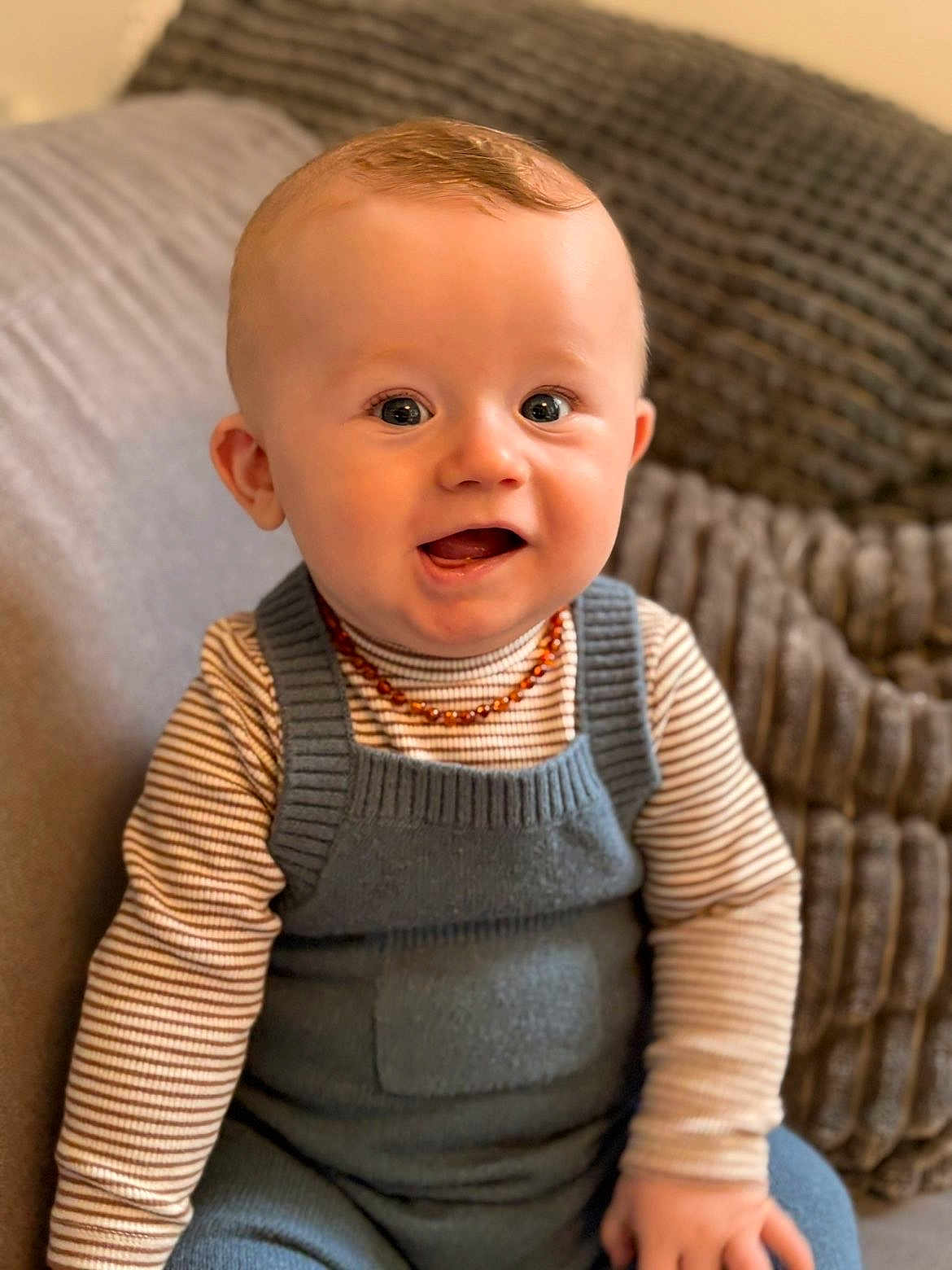Elyano a rejoint le concours — aidez-le/la à gagner de superbes lots ! baby, infant, child, smiling, portrait, sitting, striped_shirt, knit_overall, amber_necklace, couch, cushion, knit_blanket, indoor, closeup, blue_eyes, chubby_cheeks, hand, hair, adorable, warm_lighting