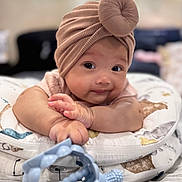 Khloe is registered to the contest to win money with this photo: baby, child, turban, hat, toy, cushion, patterned, indoor, infant, cute, smile, face, hand, reaching, lying, soft_focus, portrait, skin, expression, playful
