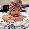 baby, child, turban, hat, toy, cushion, patterned, indoor, infant, cute, smile, face, hand, reaching, lying, soft_focus, portrait, skin, expression, playful