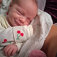 Willow is registered to the contest to win money with this photo: baby, newborn, smile, sleeping, blanket, pajamas, cherry_pattern, hand, cozy, cute, infant, soft, warm, closeup, person, child, resting, indoors, comfort, tender