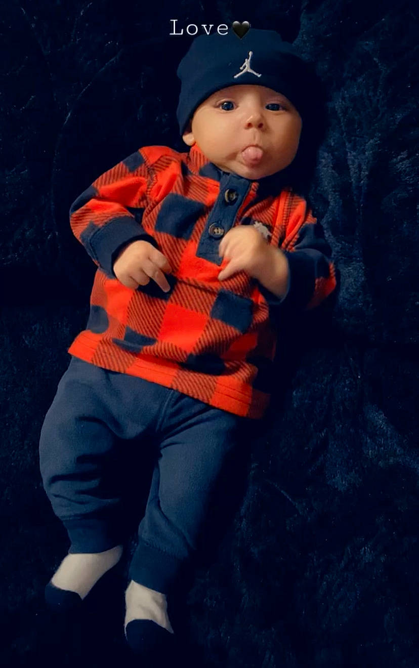 Juwan is registered to the contest to win money with this photo: baby, baby_toddler_clothing, cap, child, electric_blue, eye, face, flash_photography, gesture, head, headwear, magenta, orange, outerwear, pattern, person, personal_protective_equipment, sitting, sleeve, t_shirt