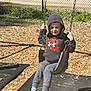 child, swing, playground, hoodie, casual_clothing, outdoor, daylight, fence, grass, wood_chips, footwear, socks, smiling, person, recreation, park, metal_chains, shoes, young_child, nature