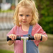 Romy participe au concours pour gagner de l'argent avec cette photo : child, toddler, blonde_hair, pony_tail, scooter, pink_shirt, denim_overalls, outdoor, greenery, blurred_background, holding, handlebars, young_child, casual_clothing, portrait, daylight, cute, curly_hair, focused, standing