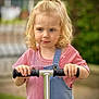 child, toddler, blonde_hair, pony_tail, scooter, pink_shirt, denim_overalls, outdoor, greenery, blurred_background, holding, handlebars, young_child, casual_clothing, portrait, daylight, cute, curly_hair, focused, standing