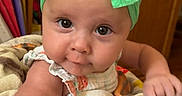 Navy is registered to the contest to win money with this photo: baby, child, green_bow, blanket, toy, face, wide_eyes, indoor, wooden_cabinet, video_call, woman, sunglasses, smile, table, colorful, person, infant, headband, arm, curious