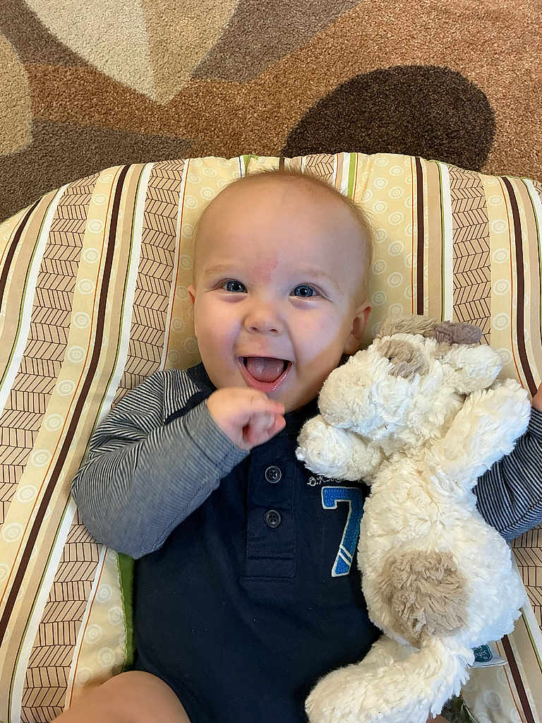 Giovanni is registered to the contest to win money with this photo: baby, child, smiling, stuffed_animal, plush_toy, cushion, striped_pattern, carpet, indoor, happy, infant, clothing, blue_onesie, face, hand, toy, cute, lying_down, comfort, playful
