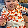 baby, child, infant, tongue, smile, pumpkin_pattern, clothing, person, indoor, furniture, crib, tattoo, hand, face, cute, happy, seated, adult, holding, blue_eyes