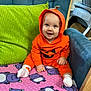 baby, child, orange_hoodie, pumpkin_costume, smiling, blue_chair, green_cushion, pink_blanket, purple_hippos, indoors, cute, happy, sitting, cozy, infant, person, footwear, white_socks, playful, portrait