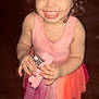 child, toddler, smiling, closed_eyes, toy, pink_dress, curly_hair, top_knot, indoor, wooden_floor, happy, person, small_hands, cute, playful, young_child, face, portrait, standing, innocence