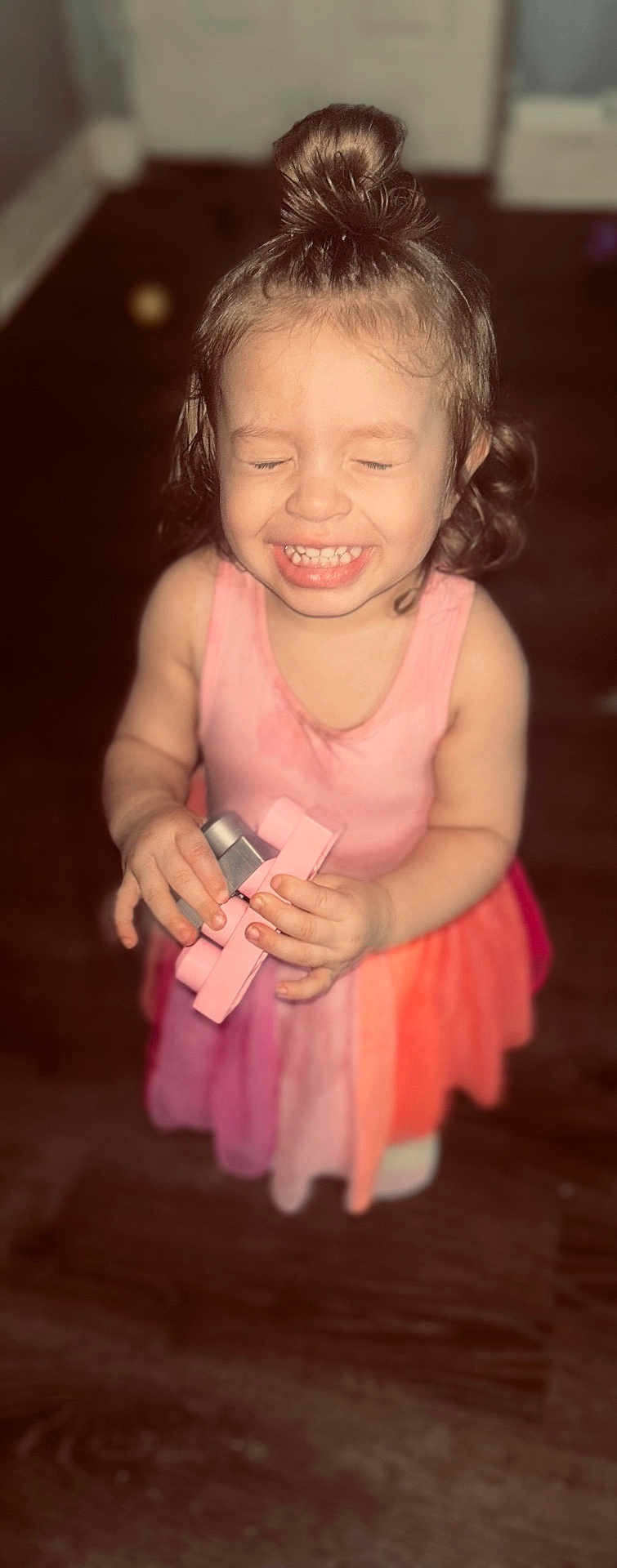 Amirah is registered to the contest to win money with this photo: child, toddler, smiling, closed_eyes, toy, pink_dress, curly_hair, top_knot, indoor, wooden_floor, happy, person, small_hands, cute, playful, young_child, face, portrait, standing, innocence