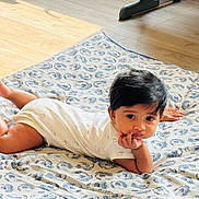 Cristiano joined the competition — help win amazing prizes! baby, child, infant, blanket, floor, wooden_floor, indoors, person, cute, resting, hand_on_chin, contemplative, patterned_blanket, exercise_equipment, home, young_child, lying_down, casual_clothing, short_sleeves, black_hair