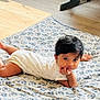 baby, child, infant, blanket, floor, wooden_floor, indoors, person, cute, resting, hand_on_chin, contemplative, patterned_blanket, exercise_equipment, home, young_child, lying_down, casual_clothing, short_sleeves, black_hair
