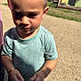 child, toddler, outdoor, sunlight, curious, holding_object, short_hair, blue_shirt, shadow, grass, building, daylight, person, young_child, casual_clothing, nature, portrait, closeup, standing, summer